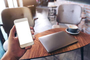 Mockup image of a man's hands holding white mobile phone with blank desktop screen , laptop and coffee cup in modern loft cafe