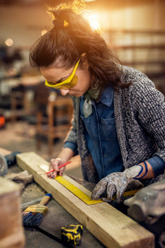 Close up view of hardworking focused professional serious carpenter woman holding ruler and pencil while making marks on the wood at the table in the fabric workshop.