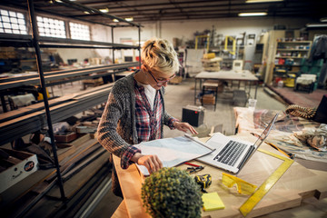 Close up view of charming smiling motivated short hair attractive middle aged industrial female engineer with eyeglasses working with blueprints and laptop in the workshop.