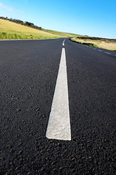grey asphalt country road with strong white lines in the middle of the road, the road has a camber road and lines big in the foreground leading to small in the distance, green rolling hills on sides