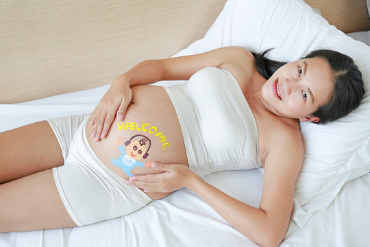 Portrait of pregnant woman belly with painted welcome happy baby and headphones lying on the bed.