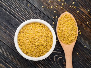 White bowl  and wooden spoon with raw bulgur on dark brown wooden background. Top view, copy space for text.