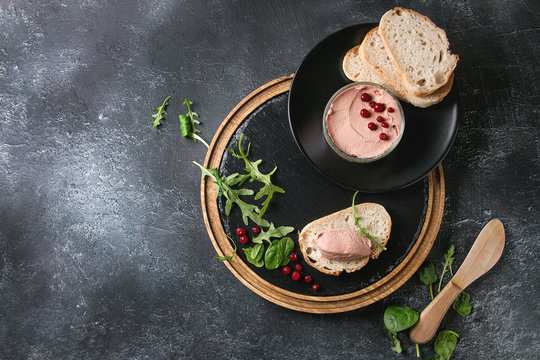 Chicken homemade liver paste or pate with sliced whole grain bread, knife, cranberries, green salad served in glass jar on wooden slate serving board over black texture background. Top view, space
