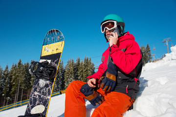 Low angle shot of a snowboarder resting sitting on the slope talking on the phone technology...