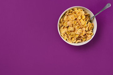 Bowl with corn flakes and spoon on purple background, top view