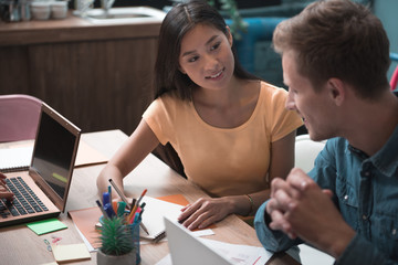 Portrait of cheerful female speaking with side view man while writing in copybook. They sitting at table. Startup concept