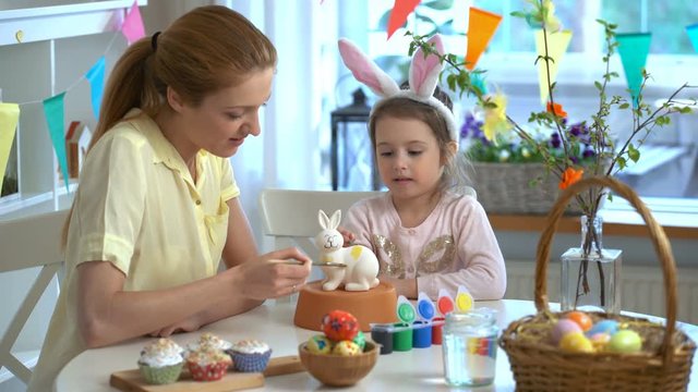 Happy Easter! Mother and her little daughter with bunny ears painting Easter Bunny at the festive table with basket and eggs