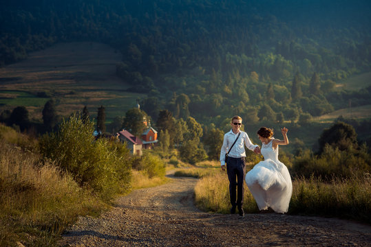 Wedding shot of the joyful attractive just married in sunglasses dancing o nthe road at the background of the mountains during the sunset.