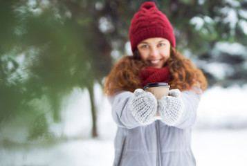 Cup of coffee in the hands of nice girl. Outdoor close up portrait of young beautiful girl with hot cup of coffee. Girl dressed in warm winter clothes and a hat posing in a winter forest.