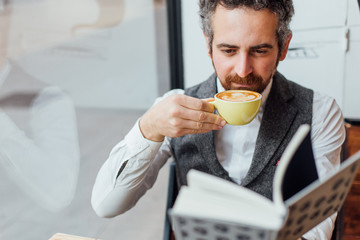 Middle aged man jewish nationality spends afternoon or morning in trendy or hipster coffee shop, sips on coffee while reading book. Competely immersed and interested in content of article
