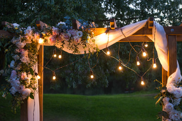 Beautiful place made with wooden square and floral decorations for outside wedding ceremony in wood. Wedding settings with electric lamp  bulbs at scenic place at night. Horizontal color photography.