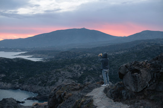 Unrecognizable man in hoodie stands on top of hiking trail on mountain, overlooks amazing beautiful scenery with sunset, makes photo on smartphone camera, concept lifestyle travel blogger