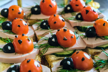 Smiling sandwiches with edible ladybugs from tomatos and olives, as well as bread, cheese, cucumber, sausage, dill. Perspective view with selective focus.