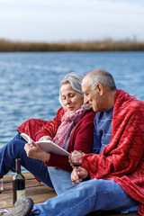 Fototapeta premium Happy senior couple enjoying time together reading a book by the lake wrap around in a red blanket.