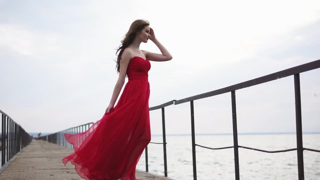 a young and pretty woman in an alat dress is standing by the river, her dress is fluttering under the streams of the wind, the lady touches her curly hair