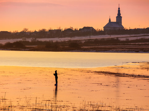 undefined person skating on the frozen lake in evening. beautiful winter countryside scenery. village and church in a far distance