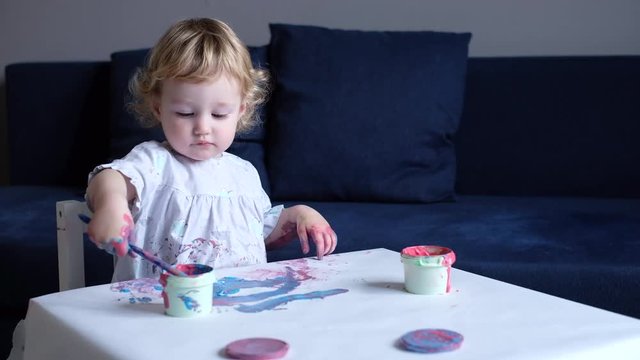 Toddler girl painting with paintbrush and colorful paints on white paper at the table