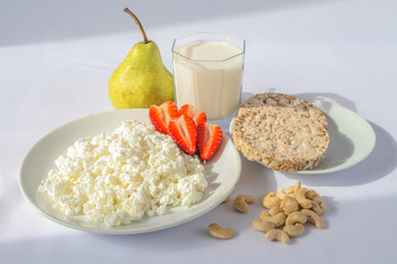 White porcelain plate with cheese and ripe strawberries, cashews and transparent glass cup with milk. green pears and crackers. White background