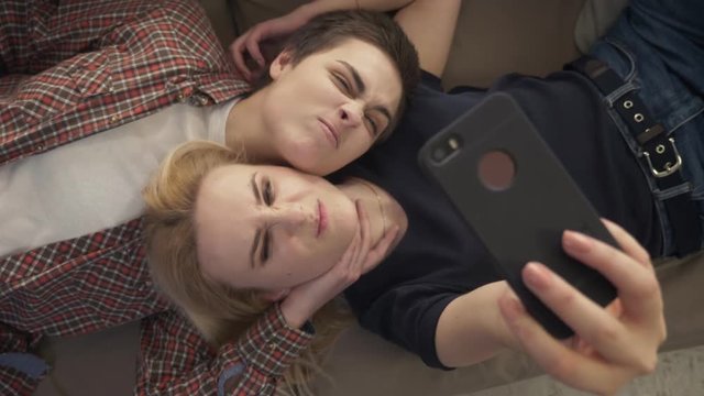 Two young girls lie on the couch, do selfie on a smartphone, grimace, make funny faces, top shot 60 fps