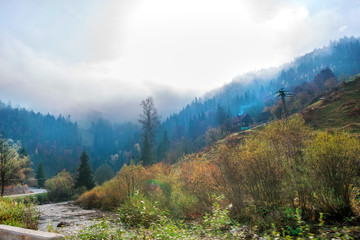 Beautiful morning on highway in mountains. Thick fog around highest mountain tops. Small river with rocks next to road. Colorful and spooky autumn background