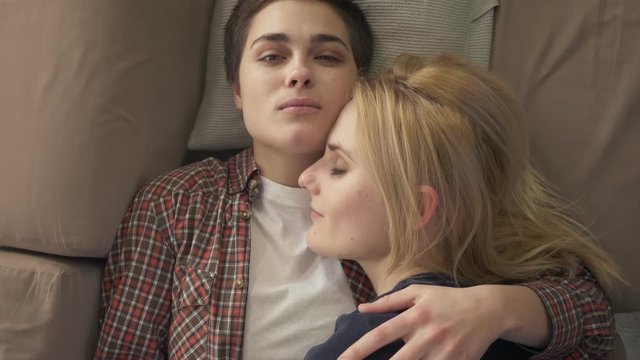Two young lesbian girls lie on the couch, hug, cuddle, sleep, girl with short hair looks at the camera, lgbt family concept, top shot 60 fps