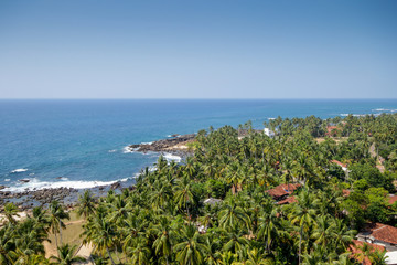 Fototapeta na wymiar Sri Lanka. Dondra. Top view from the lighthouse to the turquoise sea (ocean), rocky beach, sandy beach, green palms, red tiled roofs of houses and boats on the jig.