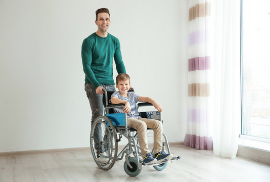 Young man taking care of little boy in wheelchair indoors