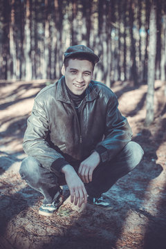 A guy in a leather jacket and cap sits on a road in a pine forest.
