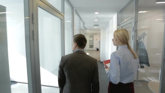 The young female HR manager is showing the office of financial company to the new male employee. The businessman and woman with red folder are going throught the long corridor with glass walls of
