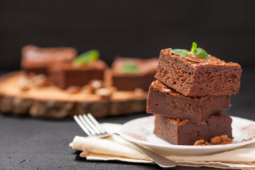 Chocolate brownie square pieces in stack on white plate with walnuts, decorated with mint leaves and cocoa on black background. Delicious dessert. Dark mood. Close up photography. Selective focus
