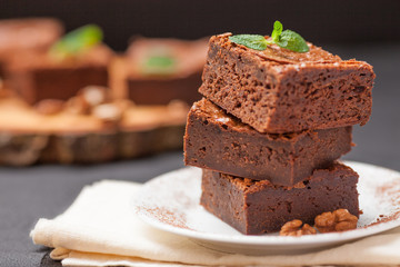 Chocolate brownie square pieces in stack on white plate with walnuts, decorated with mint leaves and cocoa on black background. Delicious dessert. Dark mood. Close up photography. Selective focus
