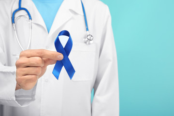 Doctor holding blue ribbon on color background, closeup. Prostate cancer awareness