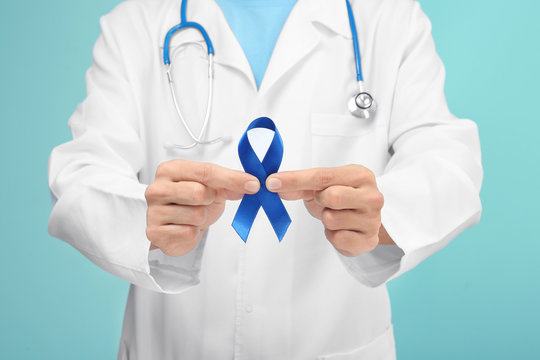 Doctor holding blue ribbon on color background, closeup. Prostate cancer awareness