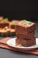 Chocolate brownie square pieces in stack on white plate with walnuts, decorated with mint leaves and cocoa on black background. Delicious dessert. Dark mood. Close up photography. Selective focus