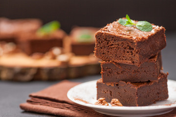 Chocolate brownie square pieces in stack on white plate with walnuts, decorated with mint leaves and cocoa on black background. Delicious dessert. Dark mood. Close up photography. Selective focus