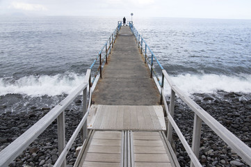 Fototapeta premium Wooden port pier with fisherman silhouette on the end, wild ocean with waves, Funchal, Madeira, Portugal