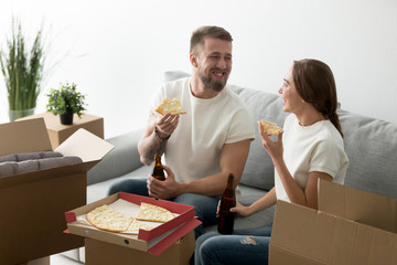 Happy homeowners eating cheese pizza drinking beer having fun together in new home glad to move in own house, young couple celebrating housewarming party taking break from packing unpacking boxes © fizkes