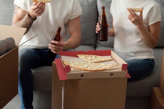 Couple eating pizza celebrating housewarming party on moving day, man and woman enjoying beer and snack sitting on sofa with unpacked boxes around, move in new home, delivery service, close up view