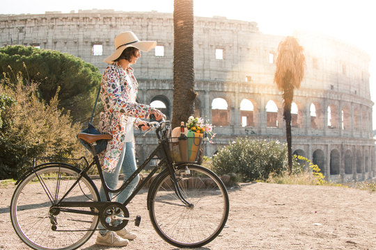 Beautiful young woman in colorful fashion dress walking alone on hill with her bike in front of colosseum in Rome at sunset with trees. Attractive tourist girl with straw hat. Lens Flare.