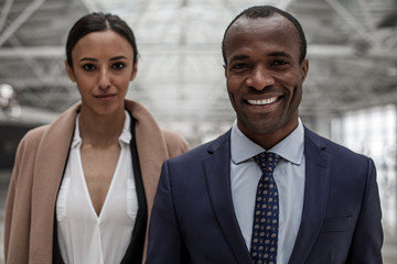 Portrait of cheerful cute young businessman and businesswoman in formal clothes are standing together and looking at camera with joy. Focus on african man