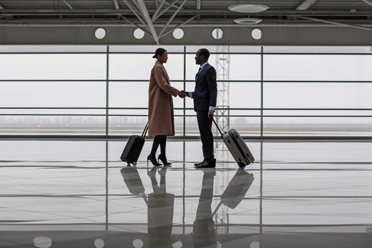 Full length of confident businessman is meeting his business partner at airport. They are standing with suitcases at lounge and shaking hands