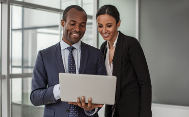 Creating new ideas. Portrait of positive young business partners wearing formal clothes are standing in office. Man is holding laptop and they are looking at screen of gadget with wide smile