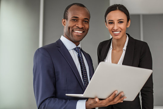 Implement new ideas. Portrait of cheerful successful young business partners wearing official clothes are standing in office. Man is keeping white laptop. They are looking at camera with joy
