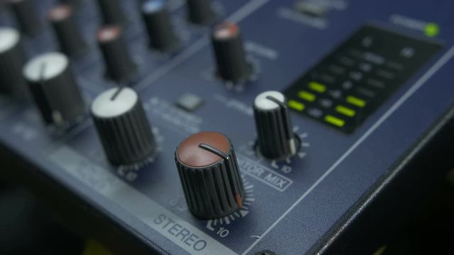 Close-up of male fingers turning fader knob on audio mixing desk. Peak level meters of an analog mixer console visible.