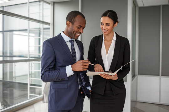 Project Management. Joyful Young Business Partners Wearing Official Clothes Are Working Together. Charming Lady Is Holding Papers While African Man Keeping Pen And Looking At Folder With Smile