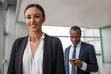 Good team. Portrait of confident successful attractive young businesswoman is standing in office and looking at camera with joy. Her colleague male is holding smartphone in background. Selective focus