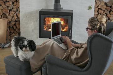 Beautiful middle-aged woman next to the fireplace relaxes in the living room and works on laptop from house. Girl in 30's surveyed for the  home bussiness project