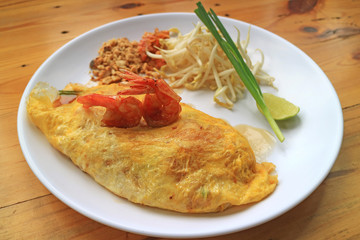 Plate of Fried Egg Wrapped Pad Thai Noodle Topped with Shrimps Served on Wooden Table