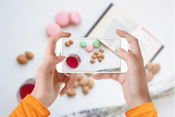 Girl taking picture of macaroons, glass of wine and book on white background with her smartphone