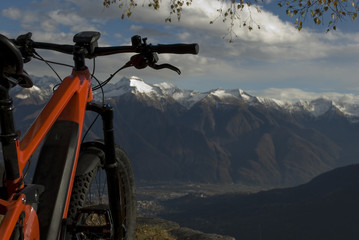 Fototapeta na wymiar ebike, e-bike, electric bicycle, high mountain, leaning against tree, detail of handlebars, wheel, saddle, display, alps landscape, snow covered tops, autumn, winter, Antrona Valley, Piedmont, Italy